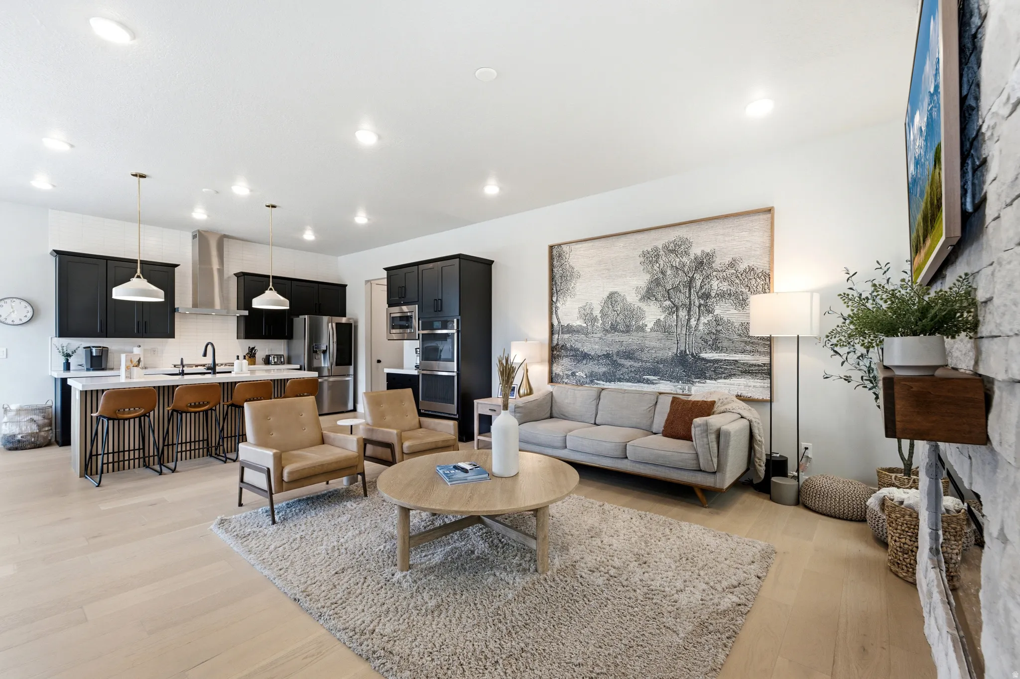 Open-concept living space featuring light wood-finish flooring, recessed lighting, and a kitchen with dark cabinetry, stainless steel appliances, and a breakfast bar island with pendant lighting