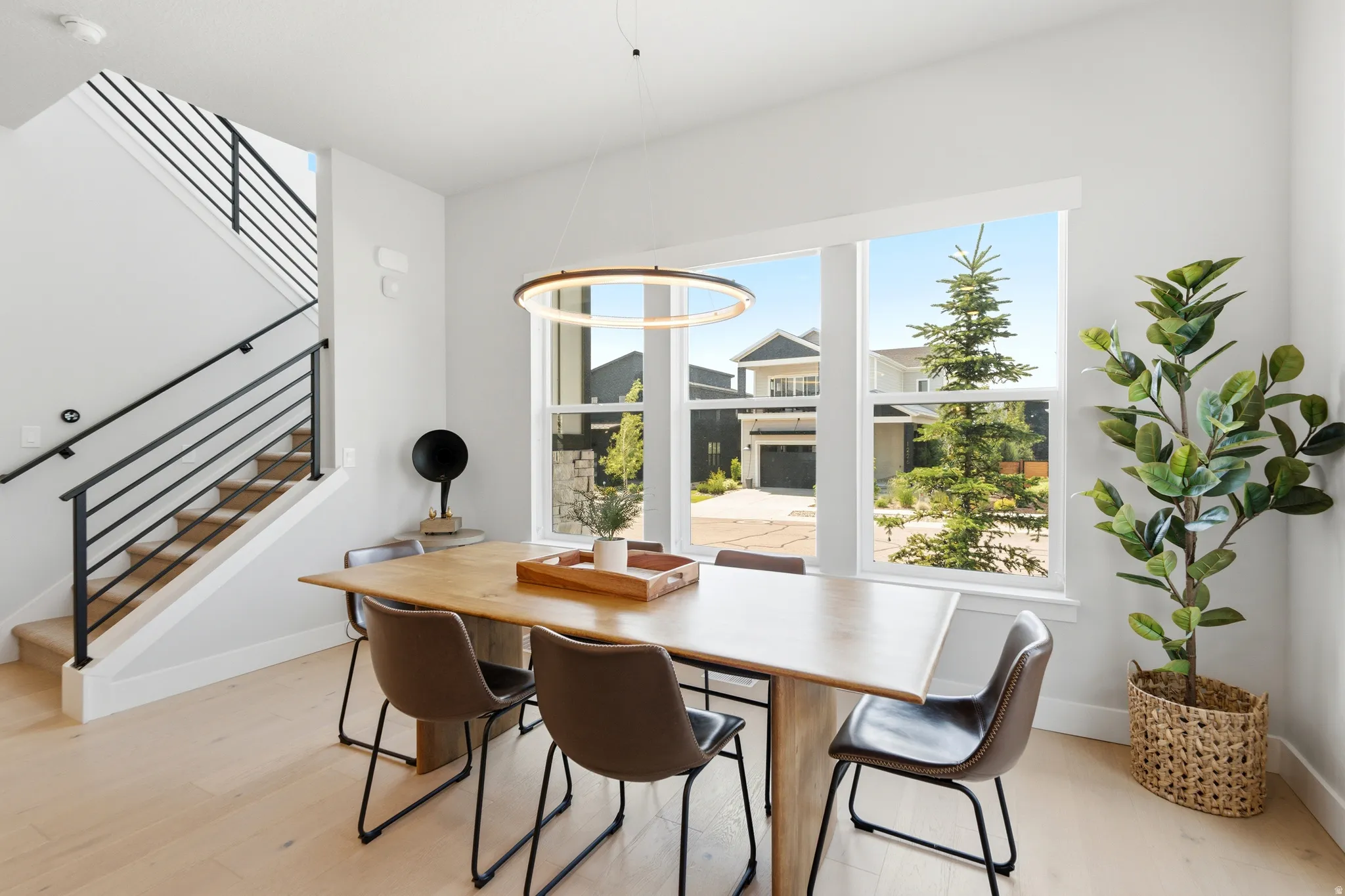 Bright dining area featuring large picture windows, wood-finish flooring, a contemporary circular pendant light, and an open staircase with a black metal railing
