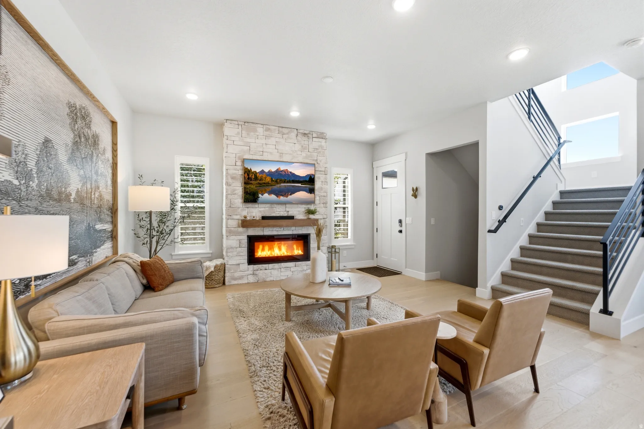 Living area featuring light wood-finish flooring, a stone-clad fireplace with a wood mantel, a white entry door with a clear glass insert, a carpeted staircase with a black metal railing, and recessed lighting