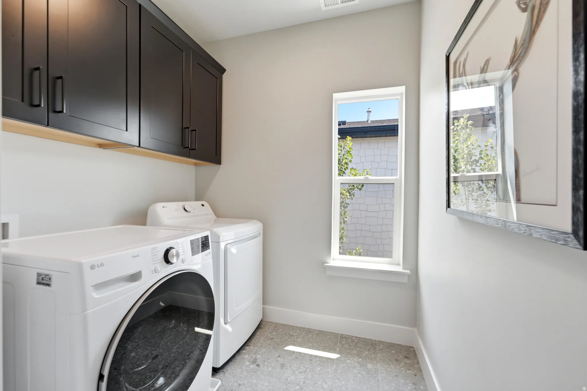 Laundry area featuring grey flooring, a single vertical window, and dark cabinetry with light wood trim