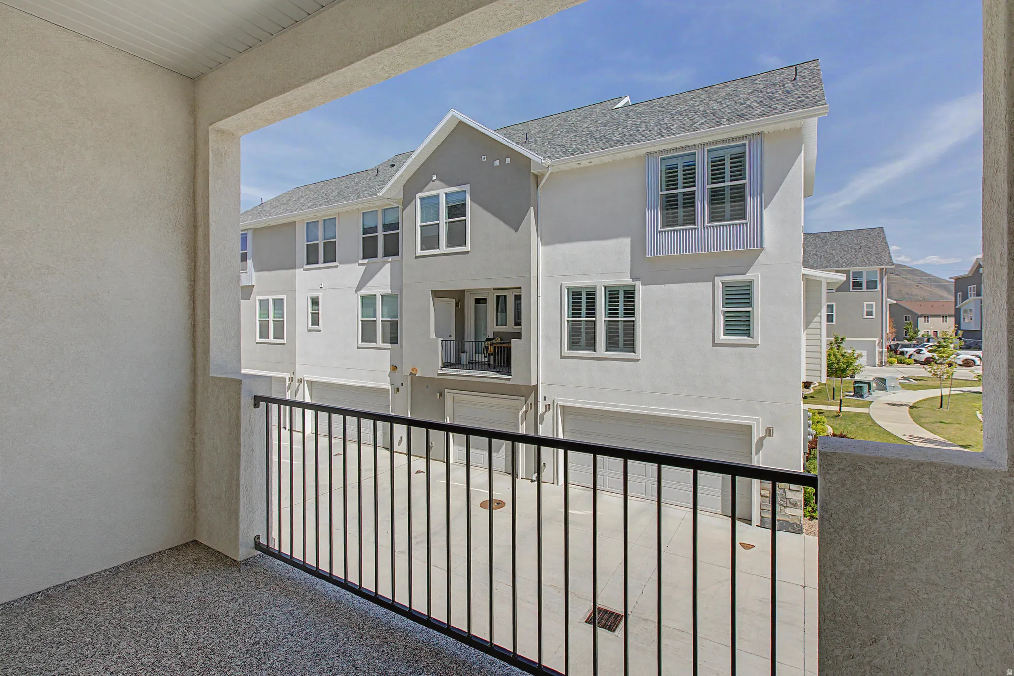 Balcony with a black metal railing and textured flooring, offering views of multi-story townhomes with light grey siding, white trim windows, and attached garages