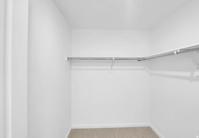 Photo of another Model Home. This home will feature Darker Cabinets and finishes. For Size and inspiration purposes only. Walk-in closet with clean white walls, light tan carpeting, and L-shaped wire shelving with integrated hanging rods