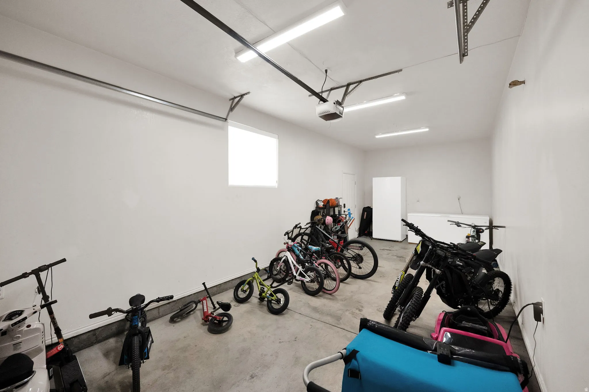 Spacious attached 3rd car garage featuring a concrete floor, bright overhead strip lighting, white painted walls, and an elevated window