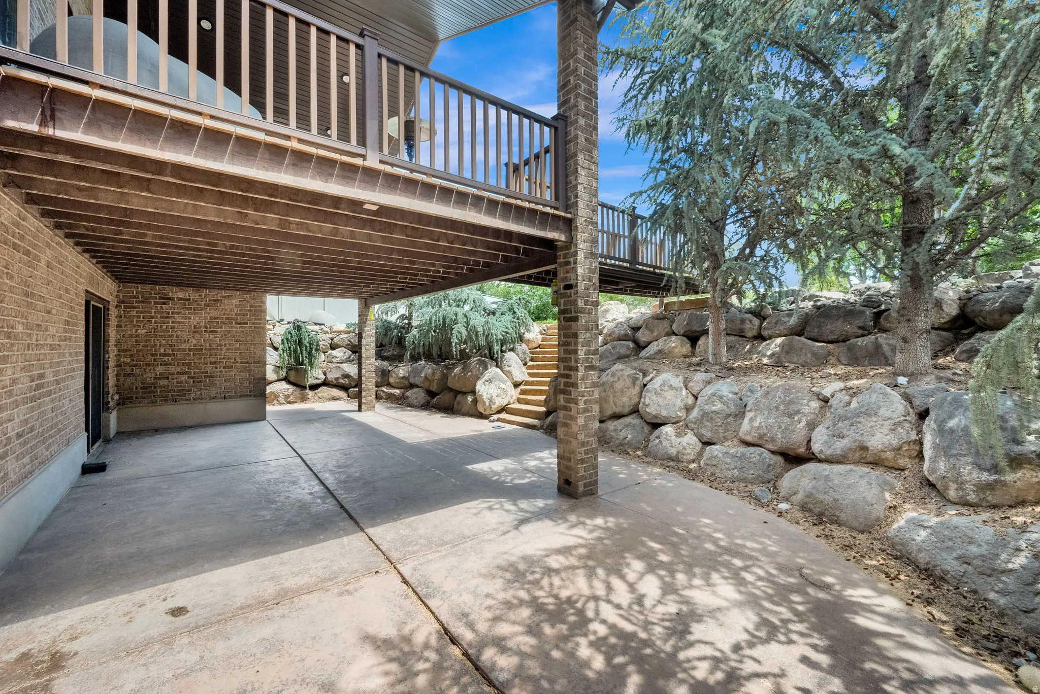 Walk out basement patio concrete patio with an overhead deck, brick exterior, and a natural rock retaining wall
