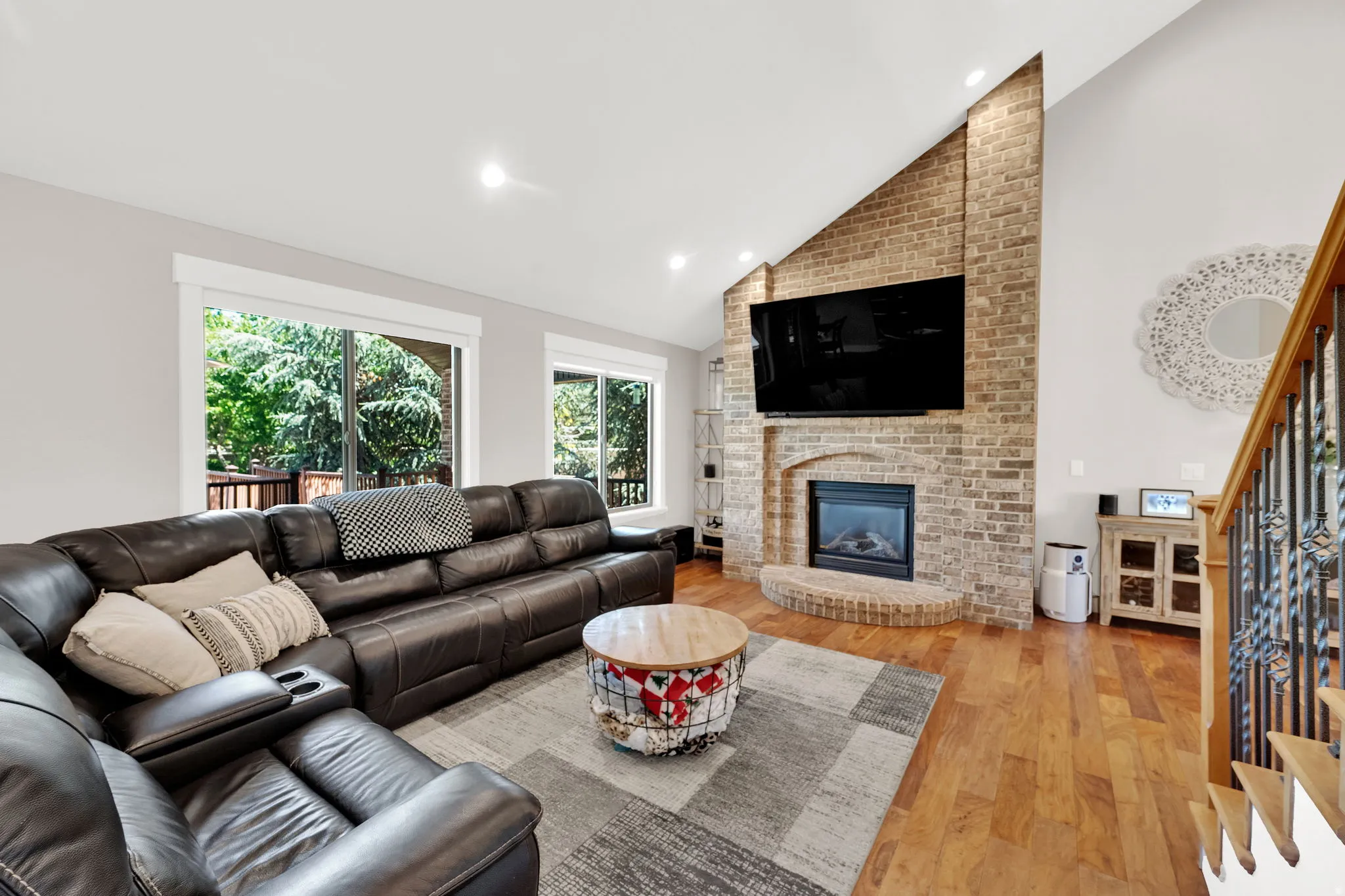 Vaulted ceiling living area featuring a brick-clad fireplace with a hearth, wood-finish flooring, and large windows overlooking exterior greenery