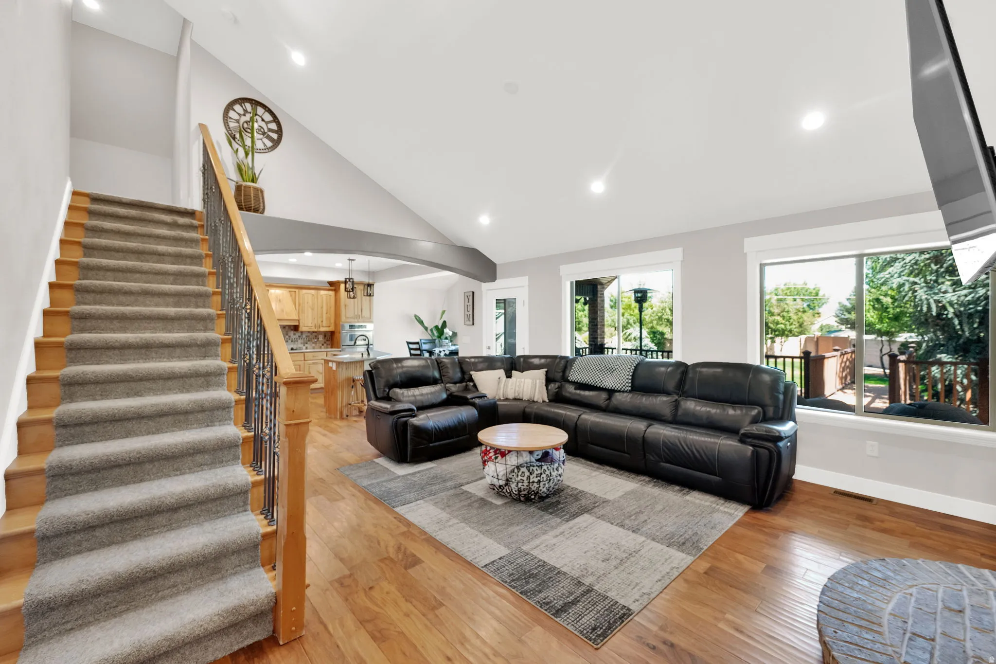 Living area featuring hardwood flooring, vaulted ceiling with recessed lighting, and large east facing windows and stairs to a bedroom.