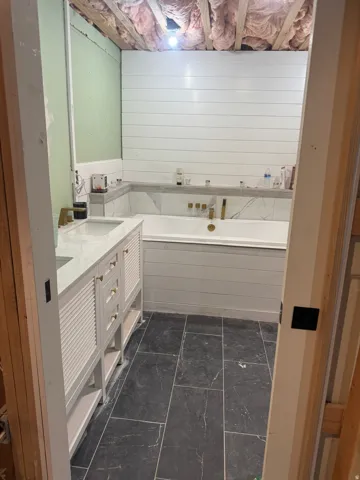 Bathroom featuring dark rectangular floor tiles, a white built-in bathtub with shiplap paneling, and a dual vanity with gold-tone hardware