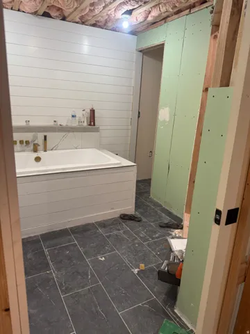 White shiplap wall paneling, built-in soaking tub with gold-finish fixtures, marble-look tile accent wall, and dark slate-look floor tiling