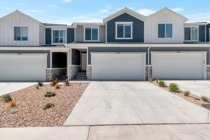 Modern townhome exterior featuring blue and white siding, stone veneer accents, individual garage doors, a covered entryway, and a low-maintenance rock landscape with mature plantings