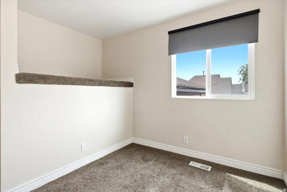 Neutral-toned room featuring a single window with a roller shade, light beige walls, and dark brown carpeting