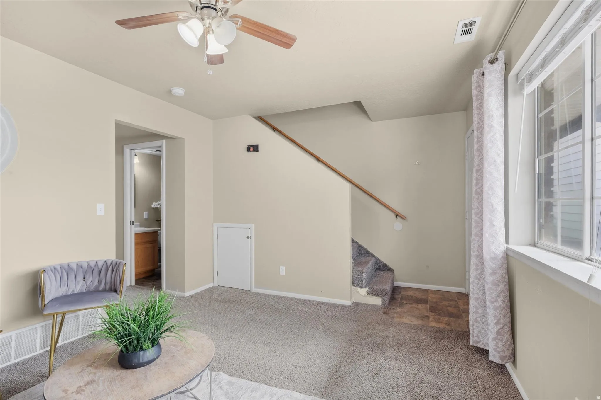 Carpeted living space featuring a ceiling fan with light fixture, large window, and staircase with wood handrail