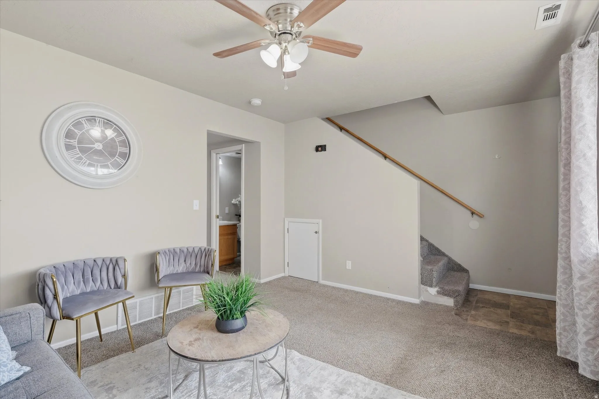 Carpeted living area featuring a ceiling fan with light fixture, neutral wall colors, and a visible staircase with wood handrail