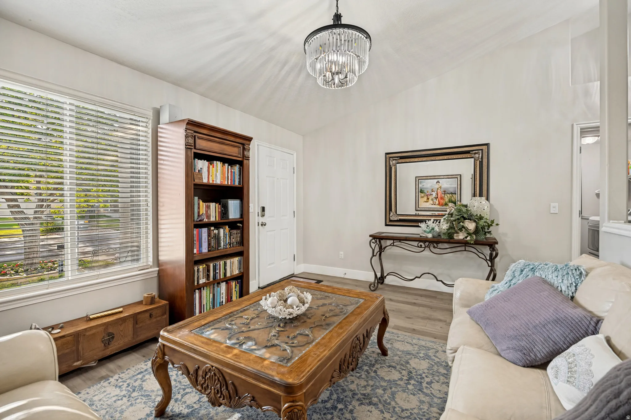 Living area featuring vaulted ceilings, wood-finish flooring, a large window with horizontal blinds, a decorative chandelier, and a built-in bookshelf