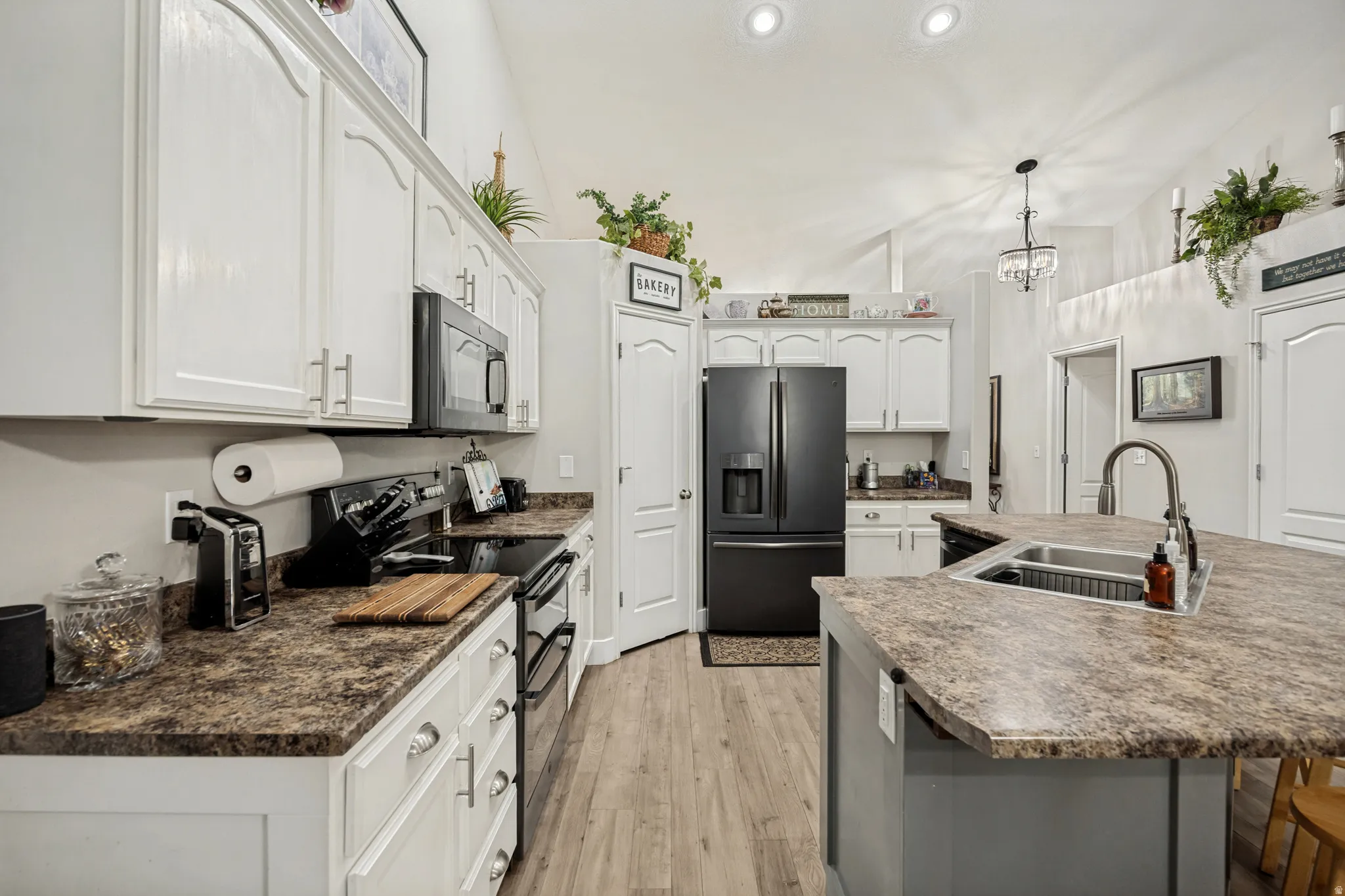 Kitchen featuring a large central island with an undermount sink, white cabinetry with silver hardware, black appliances, wood-finish flooring, and vaulted ceilings with recessed lighting