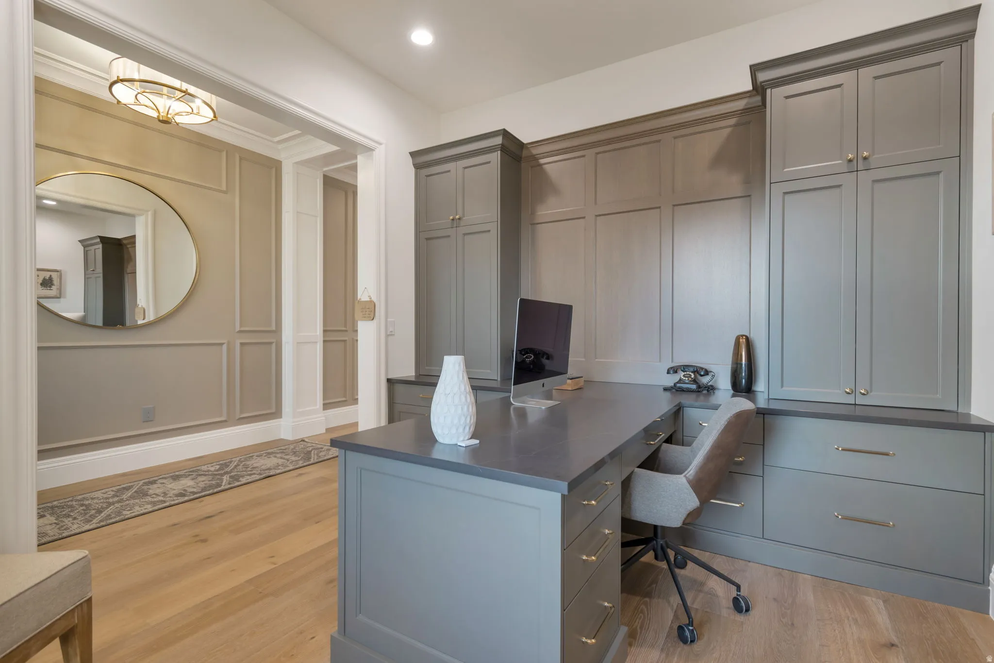 Home office featuring built-in gray cabinetry with gold-tone hardware, a dark countertop, and light wood-finish flooring