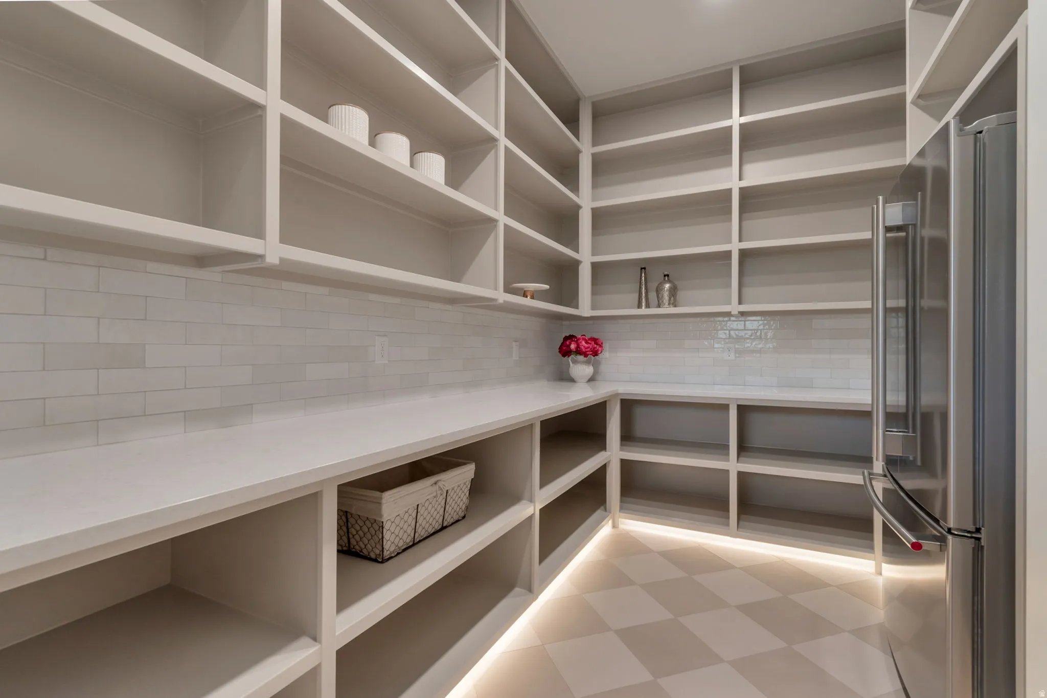 Spacious pantry featuring extensive built-in shelving, ample countertop space, a subway tile backsplash, stainless steel refrigerator, and checkerboard floor tile with under-shelf lighting