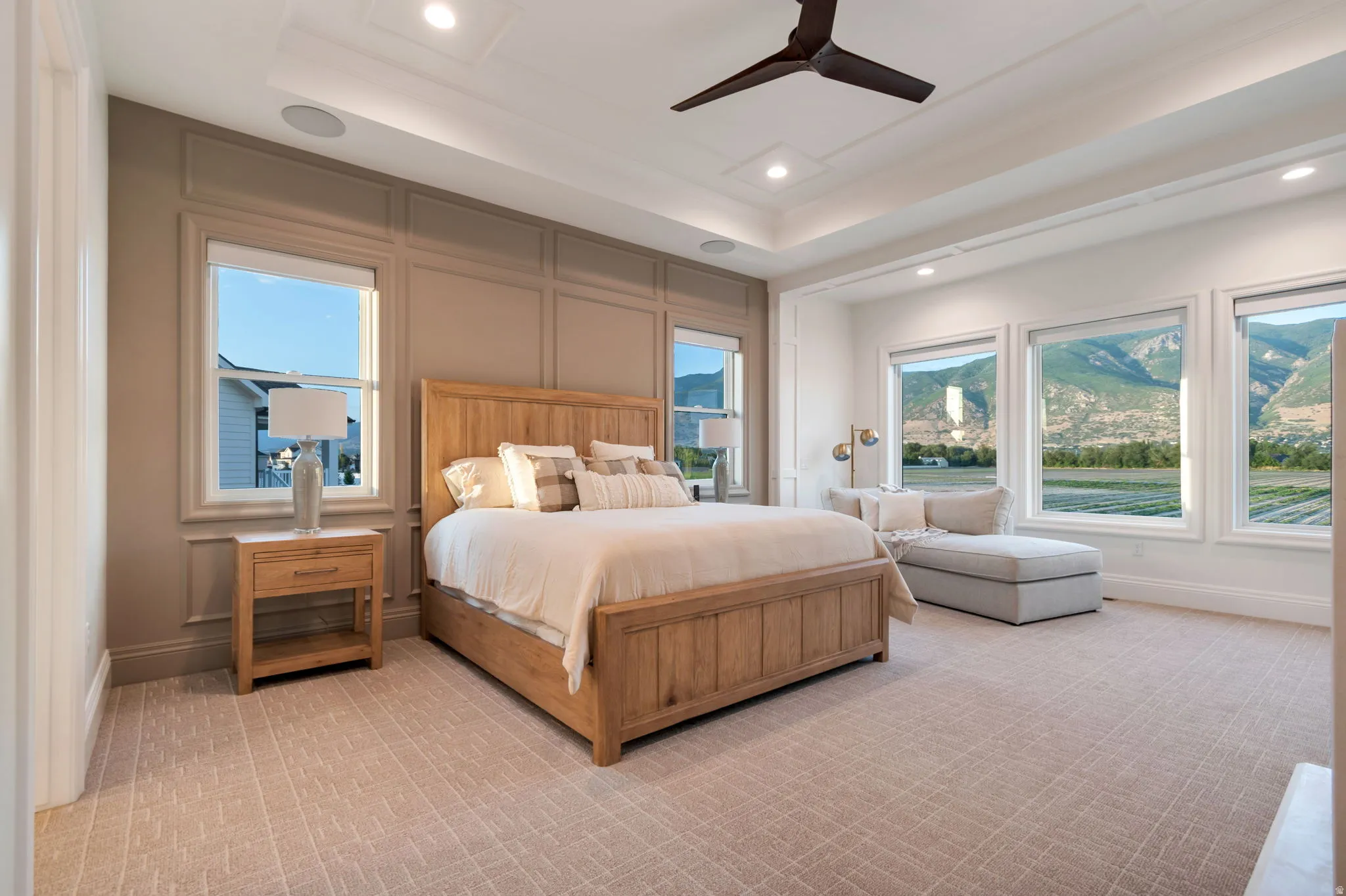Spacious bedroom featuring multiple large windows with mountain scenery, a decorative tray ceiling with recessed lighting and a ceiling fan, wainscoting on one wall, and patterned carpet flooring