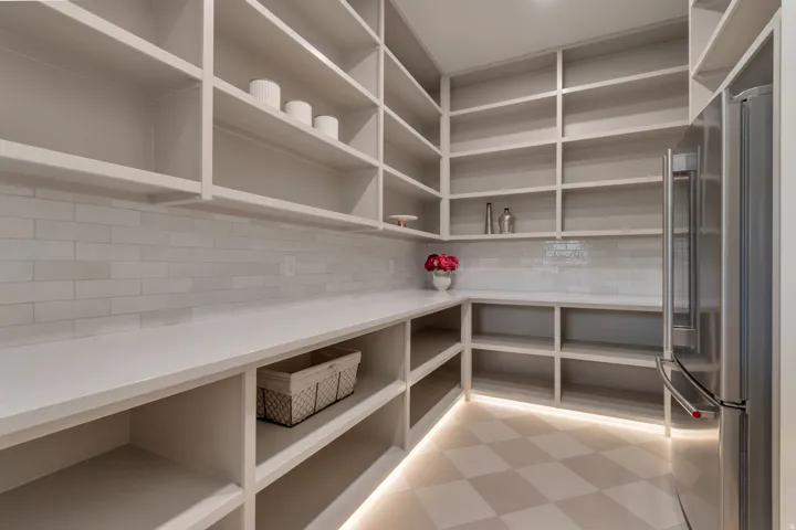 Spacious pantry featuring extensive built-in shelving, ample countertop space, a subway tile backsplash, stainless steel refrigerator, and checkerboard floor tile with under-shelf lighting