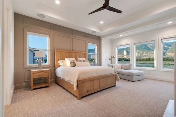 Spacious bedroom featuring multiple large windows with mountain scenery, a decorative tray ceiling with recessed lighting and a ceiling fan, wainscoting on one wall, and patterned carpet flooring