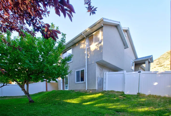 Rear exterior featuring a stucco facade, multiple windows, white vinyl fencing, and a vibrant green lawn with mature trees