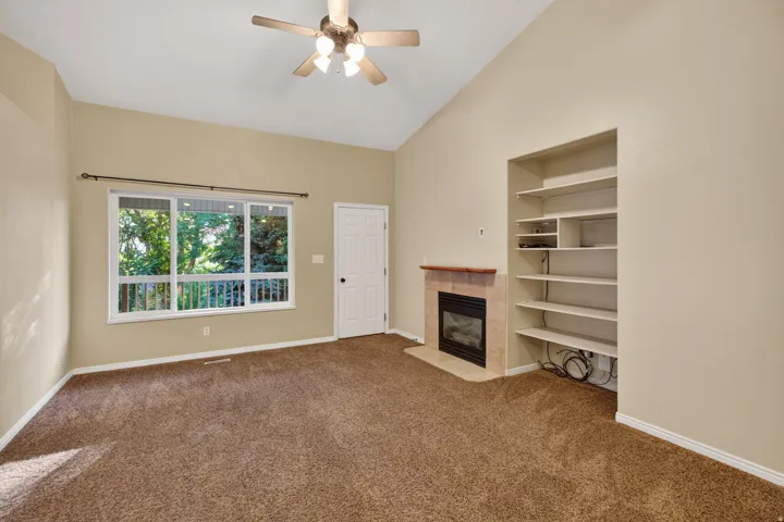 Living area featuring vaulted ceilings, a ceiling fan, broad window, built-in shelving, and a fireplace with a stone surround and wood mantel