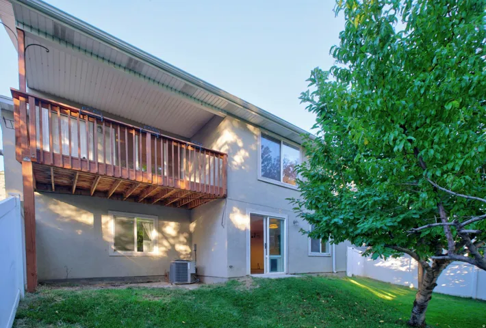 Rear exterior featuring a wood balcony, white vinyl fencing, a mature tree, and sliding glass door access to the interior