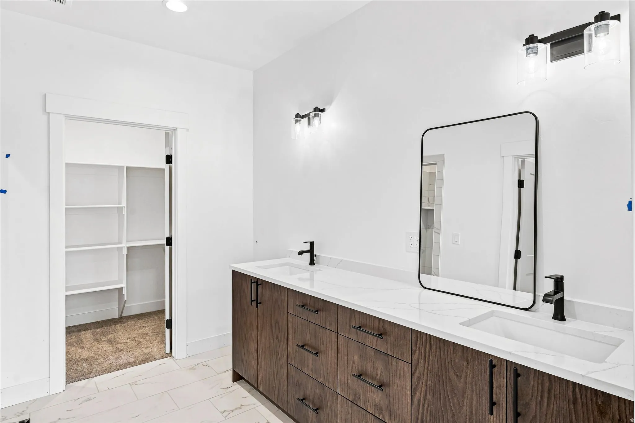 Primary bathroom featuring a double vanity with white quartz countertop, wood-finish cabinetry with multiple drawers, matte black hardware and faucetry, framed mirror with matte black trim, and recessed and sconce lighting