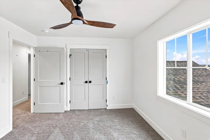 White walls, gray carpeting, a large window, a ceiling fan with wood-look blades, and white bi-fold closet doors