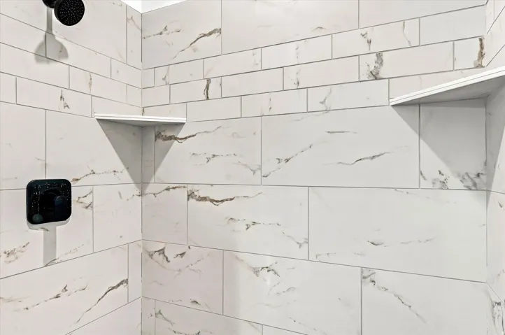 Shower with white and gray tiled walls, two built-in corner shelves, a matte black showerhead, and a matte black square-shaped control panel