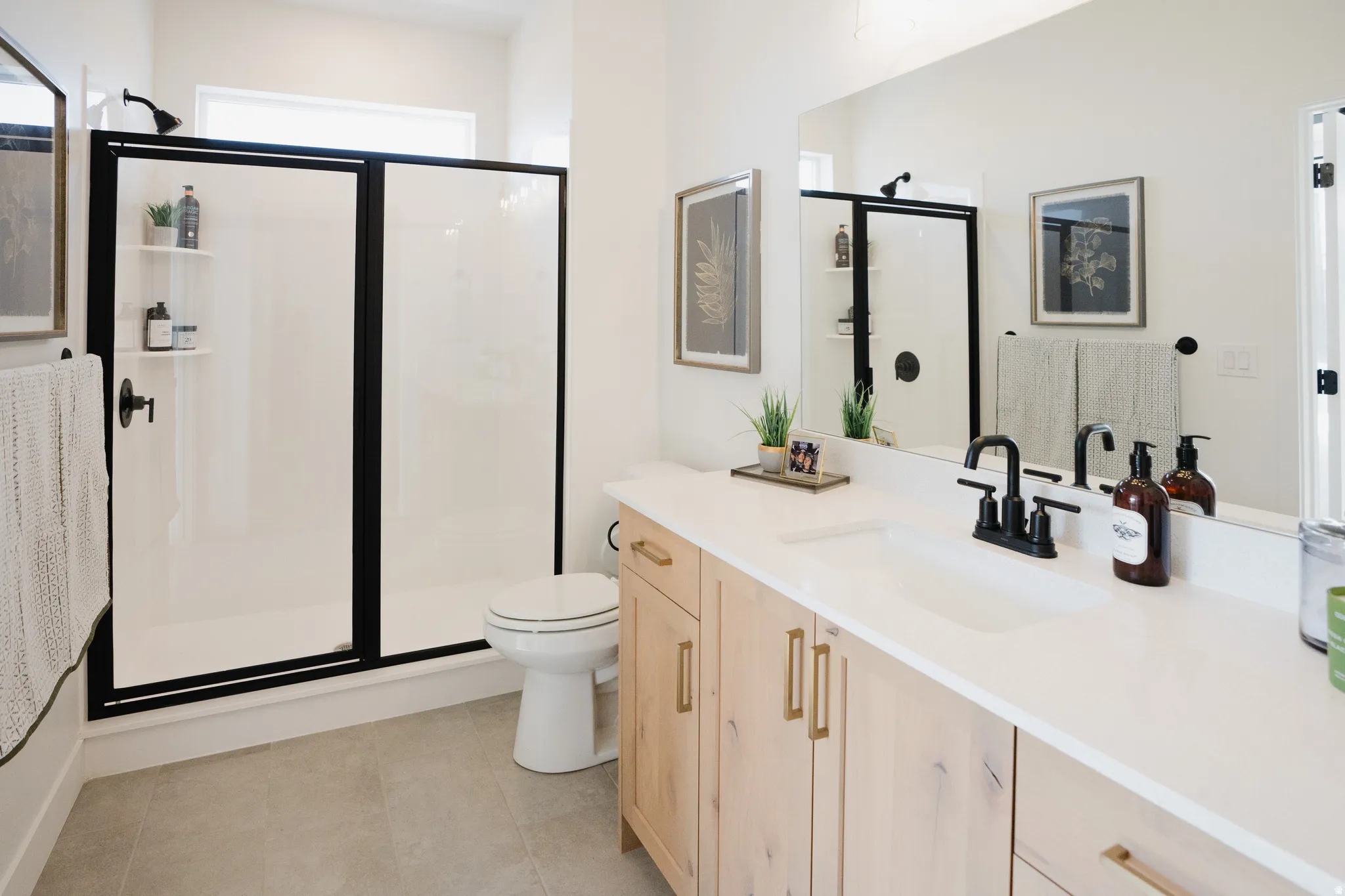 Bathroom featuring a spacious walk-in shower with black-framed glass doors, a single vanity with light wood-finish cabinetry, a white countertop, and matte black fixtures