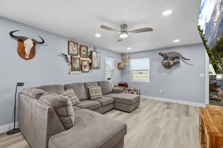 Spacious living area featuring light-toned wood-finish flooring, two large windows with horizontal blinds, and a ceiling fan with integrated lighting