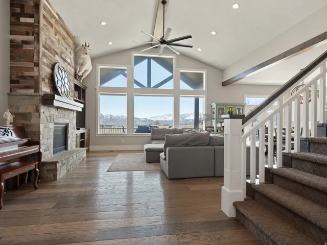 Vaulted ceiling great room featuring a stone fireplace with wood-finish mantel, multi-pane windows showcasing mountain scenery, wood-finish flooring, and a contemporary ceiling fan