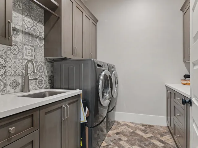 Utility space featuring patterned tile flooring, a stainless steel sink with a high-arc faucet, quartz countertops, extensive cabinetry, and decorative backsplash tiling