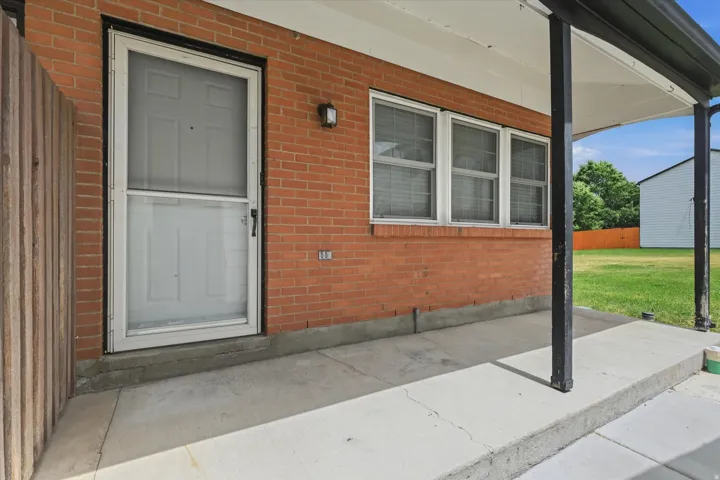 Brick exterior featuring a covered concrete patio, a white entry door with an exterior screen door, and multiple windows with blinds