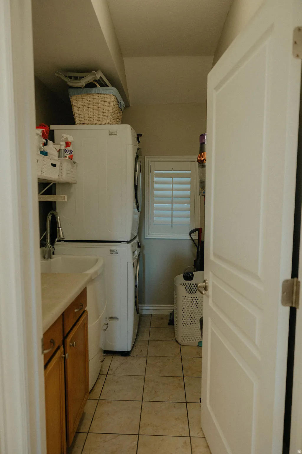 Laundry area featuring stacked appliances, tile flooring, a utility sink with a countertop, wood cabinetry, and a window with plantation shutters