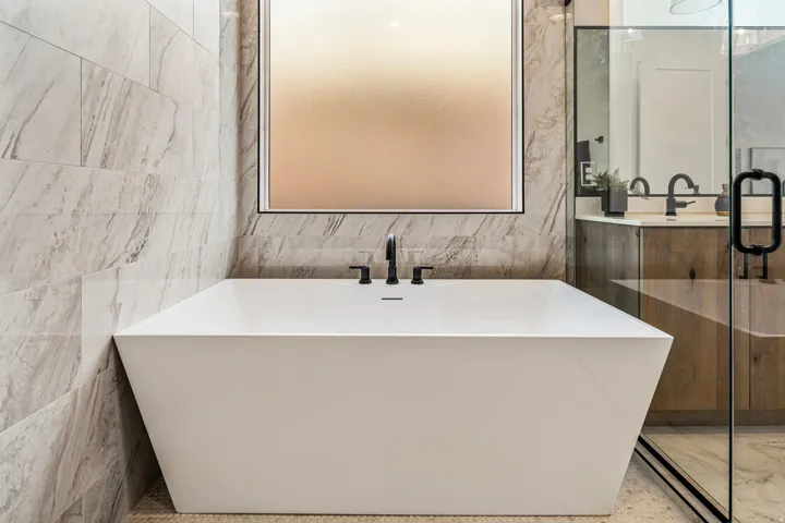 Freestanding soaking tub with matte black fixtures, set against a tiled accent wall and a frosted privacy window