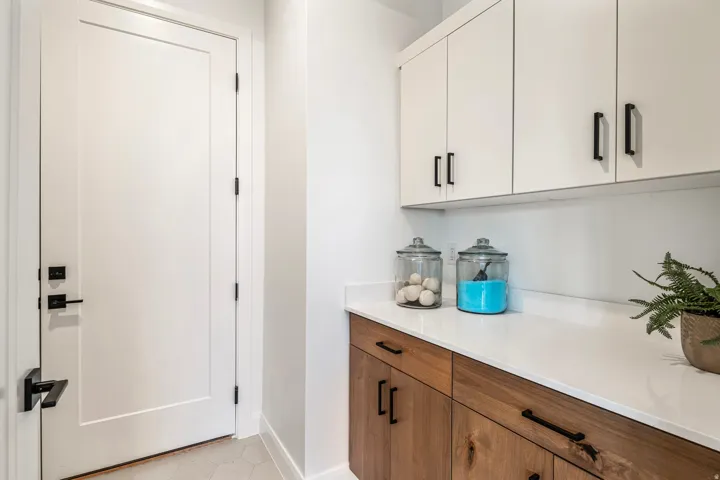 Clean-lined laundry space featuring a white flat-panel door with matte black hardware, white upper cabinetry, natural wood-finish lower cabinetry, and a light countertop