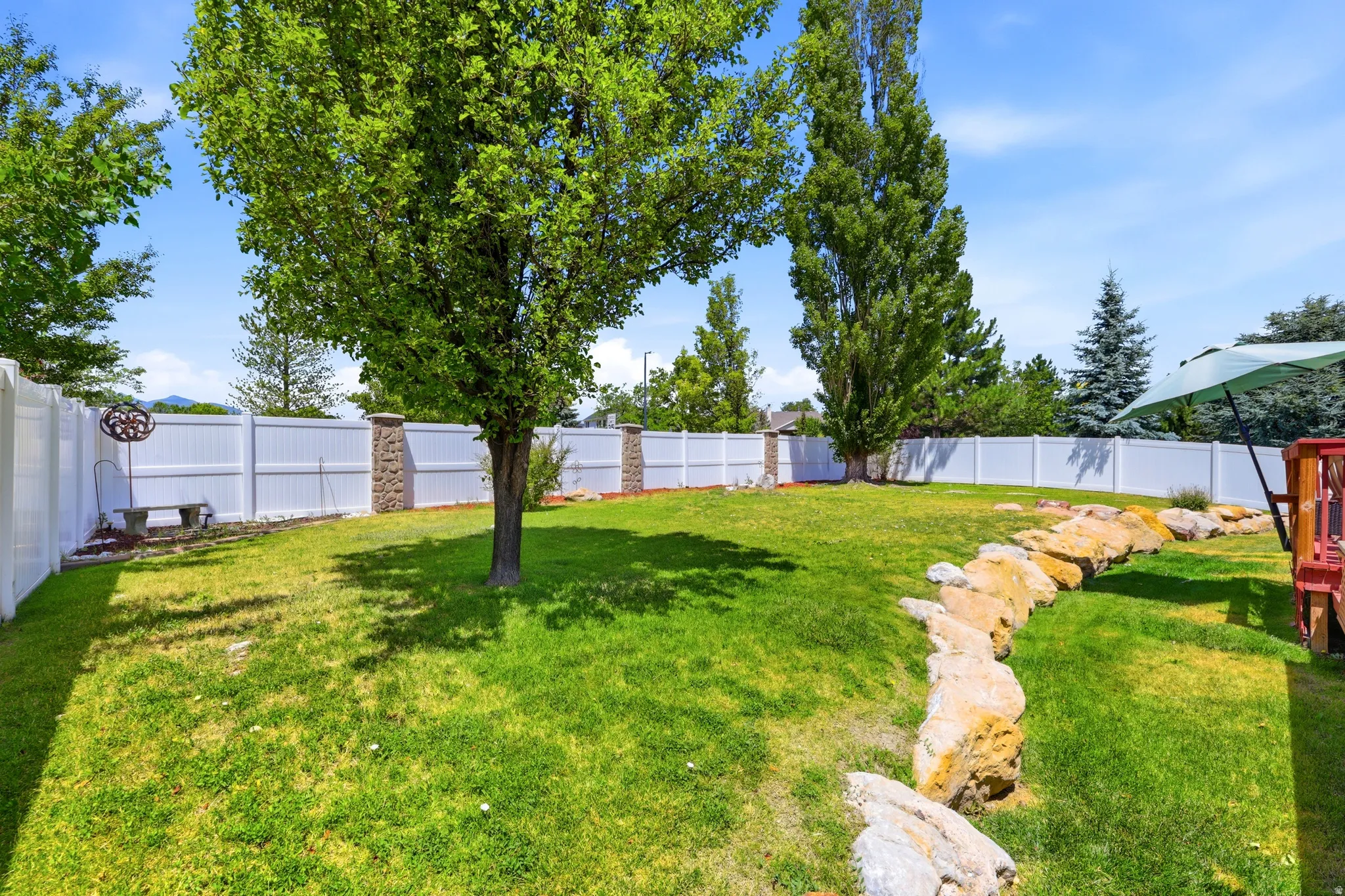 Expansive grassy backyard featuring a white privacy fence with stone pillars, mature trees, and a rock retaining wall