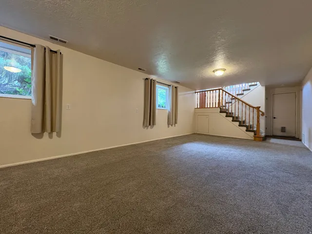 Spacious carpeted room featuring a wooden staircase with balusters, two windows with textile curtains, recessed under-stair storage, and overhead lighting
