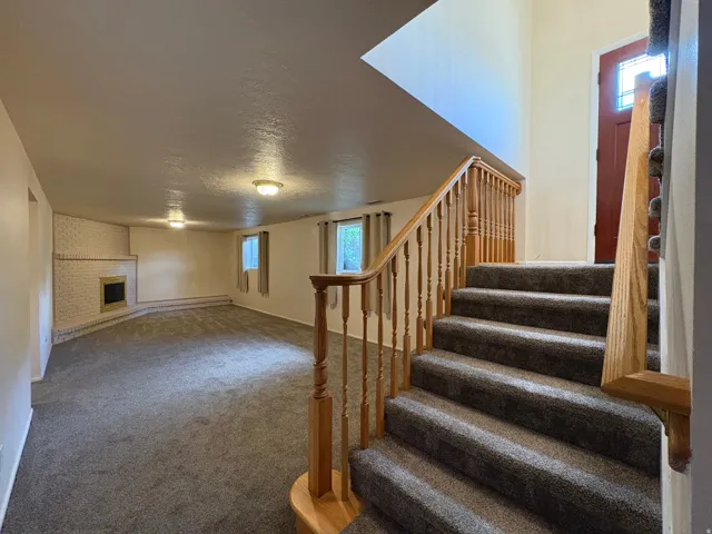 Carpeted staircase with natural wood railing leading to a spacious lower level featuring a white brick fireplace with a raised hearth, multiple windows, and overhead lighting