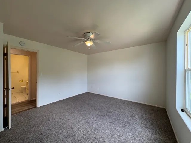 Spacious room featuring neutral carpeting, a ceiling fan with integrated lighting, and a large window providing natural illumination