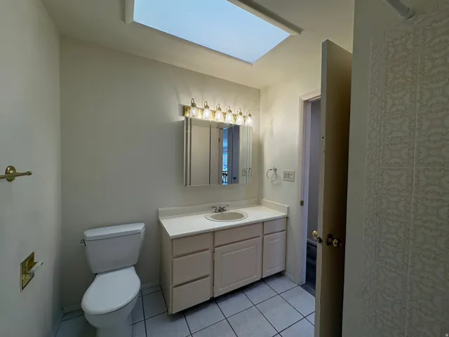 Bathroom featuring a skylight, white tile flooring, a single vanity with a white countertop and integrated sink, a mirrored medicine cabinet, and a five-light fixture