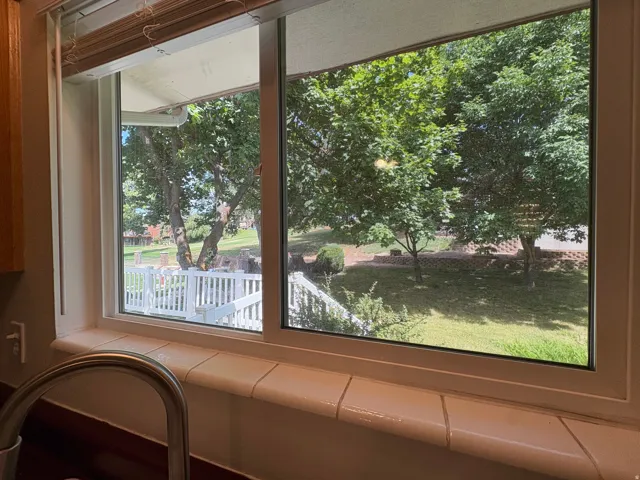 Kitchen window featuring a tiled sill, stainless steel faucet, and a backyard landscape with mature trees and a white railing