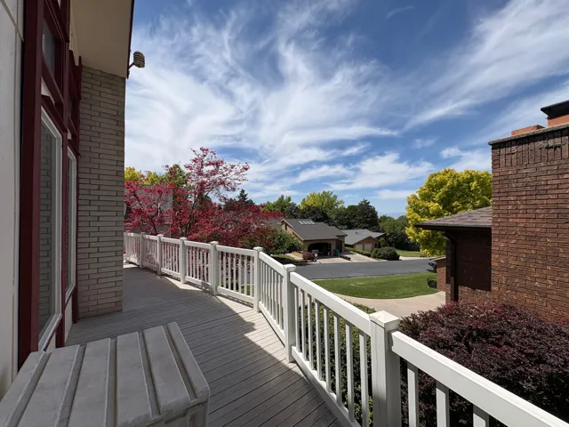 Spacious outdoor deck with composite decking and white railing, featuring a large window with red trim, light-colored brick exterior, and surrounding mature trees