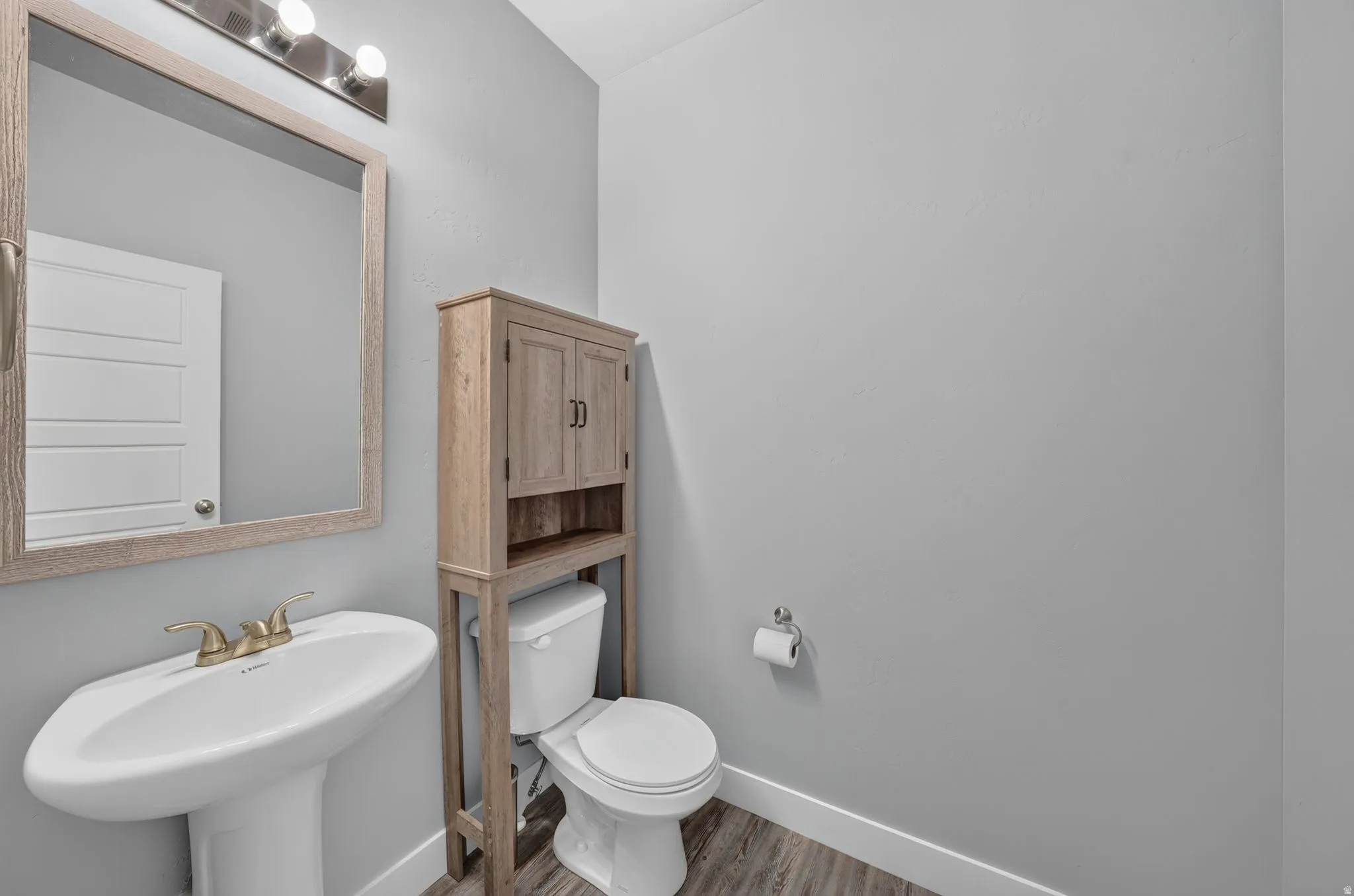 Powder room featuring a pedestal sink with brass-finish faucet, a toilet with an over-the-toilet storage cabinet, and wood-finish flooring