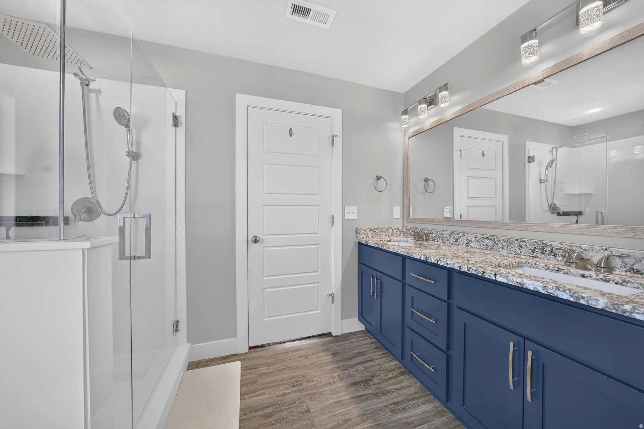 Bathroom featuring a frameless glass shower enclosure with a rain shower head and hand-held sprayer, a double vanity with navy cabinetry, a light-toned countertop, and wood-finish flooring