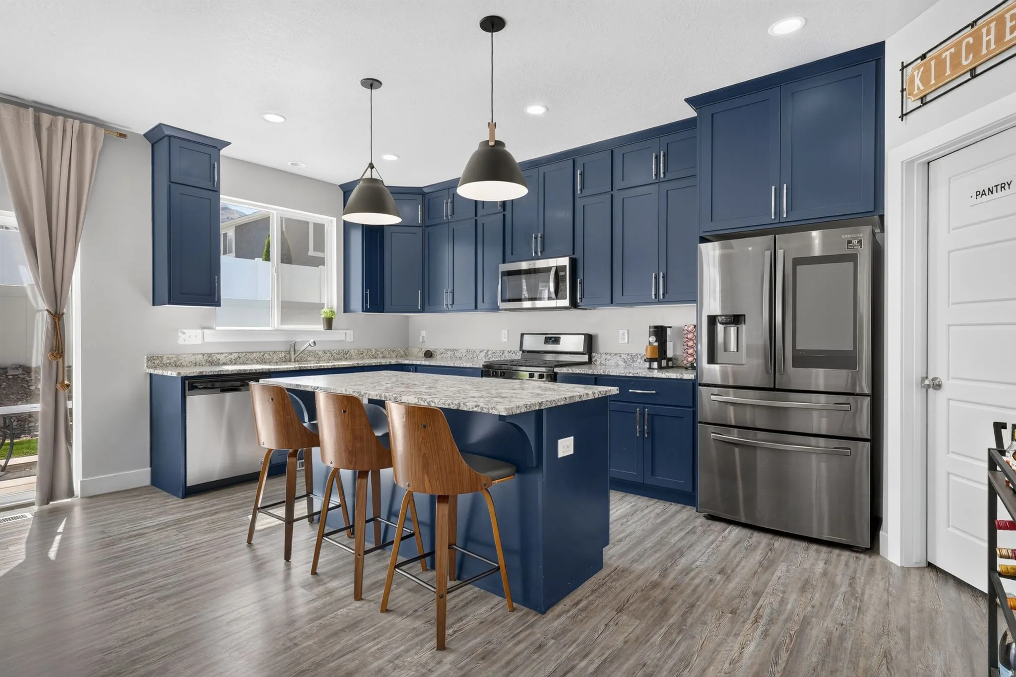 Kitchen featuring navy blue cabinetry, stainless steel appliances, light-toned granite countertops, wood-finish flooring, and pendant lighting