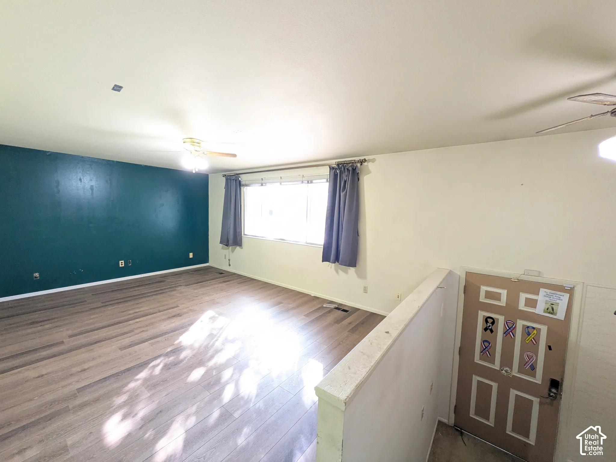 Wood-finish flooring with a contrasting accent wall, overhead ceiling fans, and a window with curtain rod