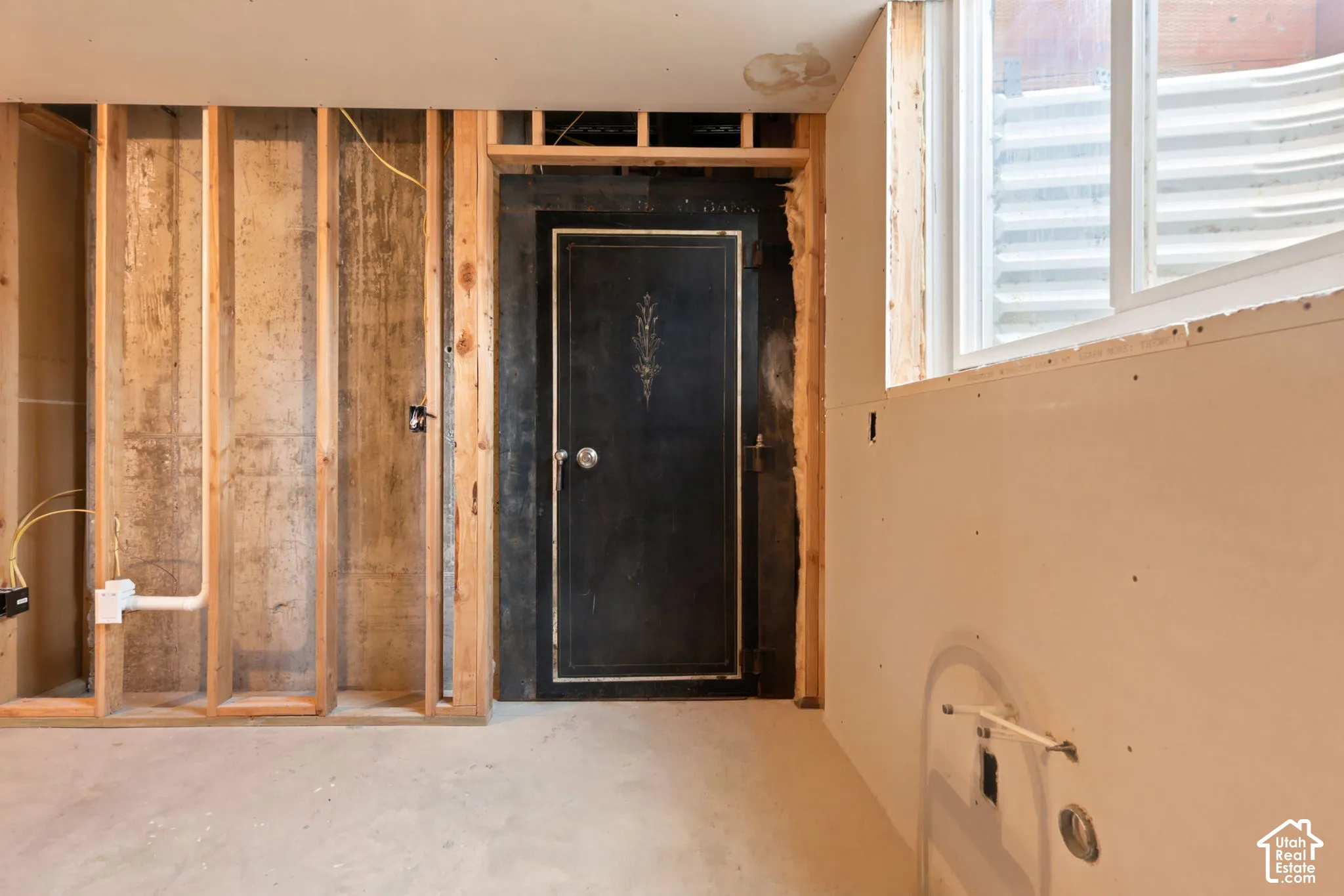 Unfinished basement space featuring a window, framed wall sections, exposed concrete foundation, and a dark, ornate built-in safe that holds nine people.