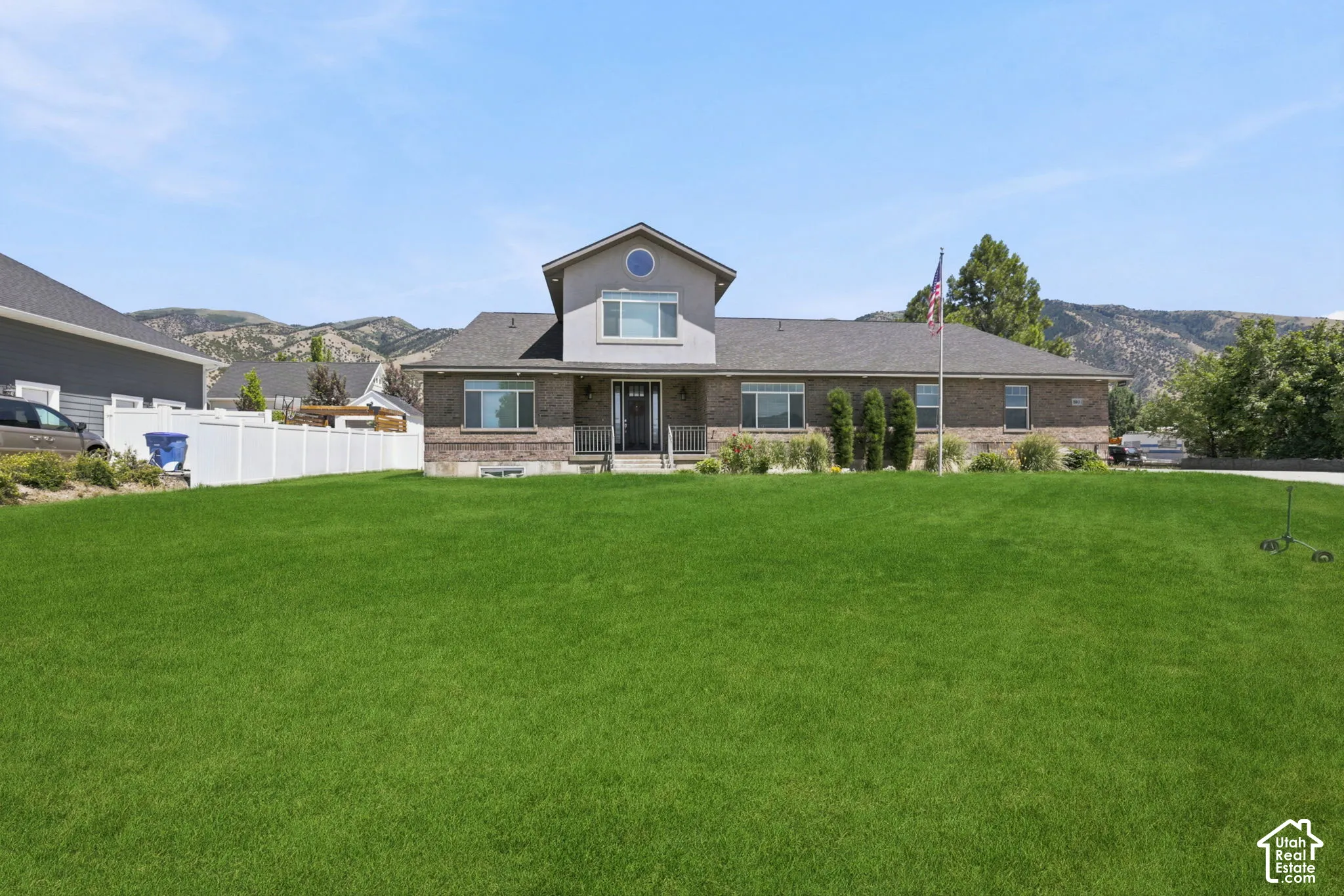 Expansive front lawn with a brick and siding exterior, a white picket fence, and a mountain backdrop  Digitally staged.